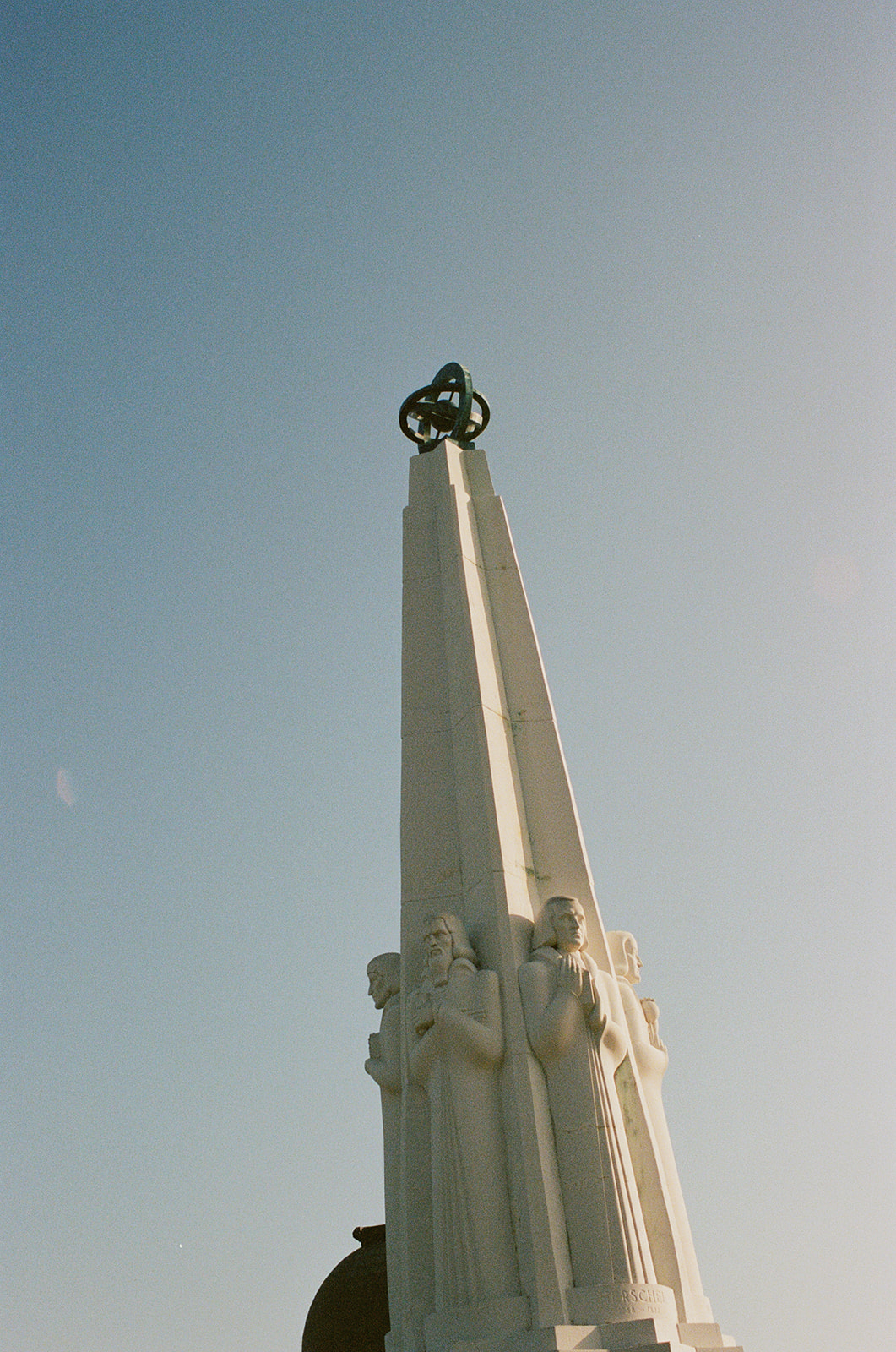 A photograph of a white Art Deco-style obelisk with a bronze decoration of some rings on top of it.